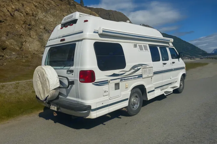 White Pleasure-Way camper van, a popular RV rental, parked on a paved road with mountains in the distance, perfect for scenic travel.. Pleasure Way Excel 2001
