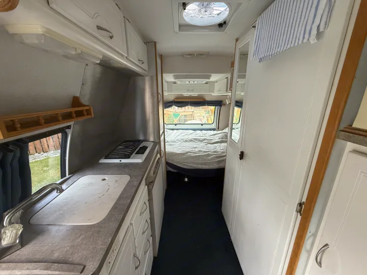 View inside an RV rental showing a compact kitchen with sink and stove, leading to a bed. Ideal for travel adventures.. Pleasure Way Excel 2001
