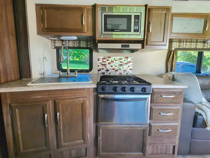 RV rental kitchen featuring a sink, microwave, Suburban stove, wood cabinets, and window offering a lush outdoor view.. Coachmen Apex 2018