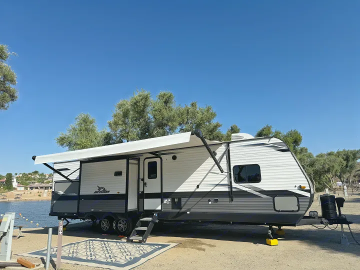 Jay Flight RV rental at a scenic lakeside campsite. The gray & white travel trailer has its awning open & outdoor rug.. Jayco Jay Flight 2022