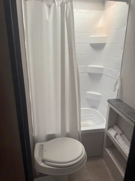 Inside an RV rental: a compact bathroom with a toilet, shower/tub combo, and a shelf stocked with fresh towels.. Forest River Palomino Puma XLE Lite 2021