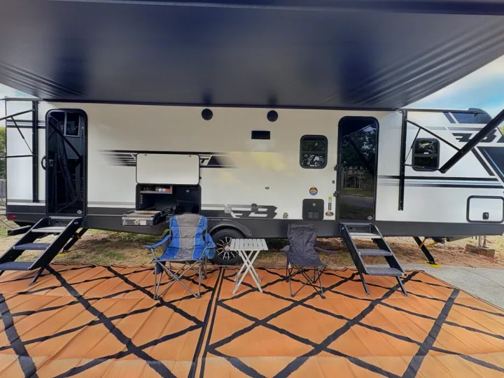 RV rental setup: Modern travel trailer with extended awning, outdoor kitchen, and camping chairs at a sunny campsite.. Keystone Bullet Crosfire 290BHS 2025