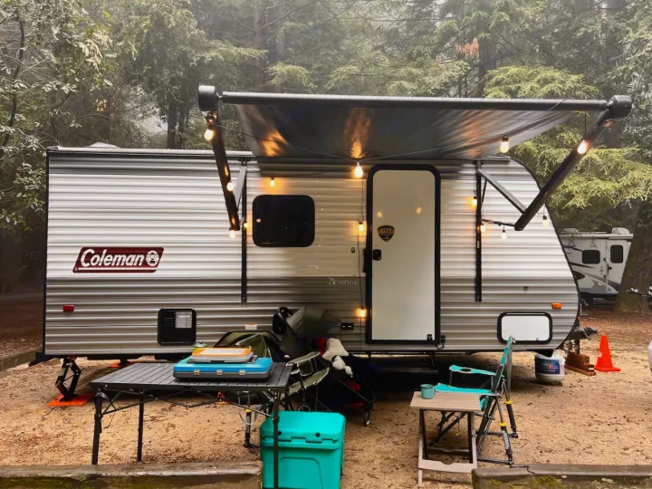This Coleman **RV rental**, with an illuminated awning, is set up at a wooded campsite with outdoor gear. Perfect for a cozy getaway.. Dutchmen Coleman Lantern LT 2023