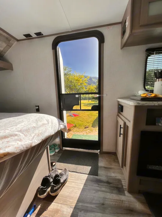 Step into this bright RV rental, featuring a cozy bed, kitchen nook, and views of a sunny, tree-lined landscape.. Dutchmen Coleman Lantern LT 2023