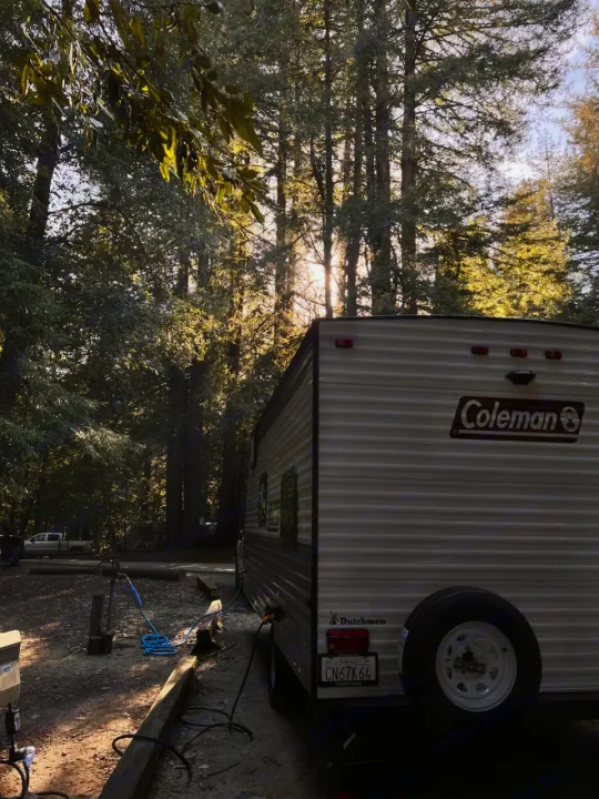 RV rental parked at a serene forest campsite with sunlight filtering through tall trees.. Dutchmen Coleman Lantern LT 2023