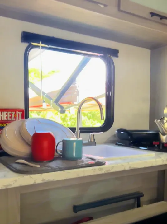 This RV rental kitchen features a sink, drying dishes, stovetop, and window view, ideal for compact travel.. Dutchmen Coleman Lantern LT 2023