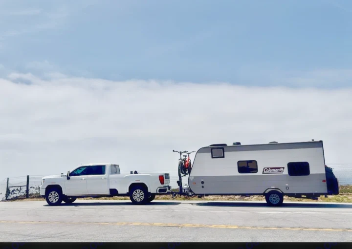 A white truck tows an RV rental Coleman travel trailer with a bike, ready for outdoor adventures under a cloudy sky.. Dutchmen Coleman Lantern LT 2023