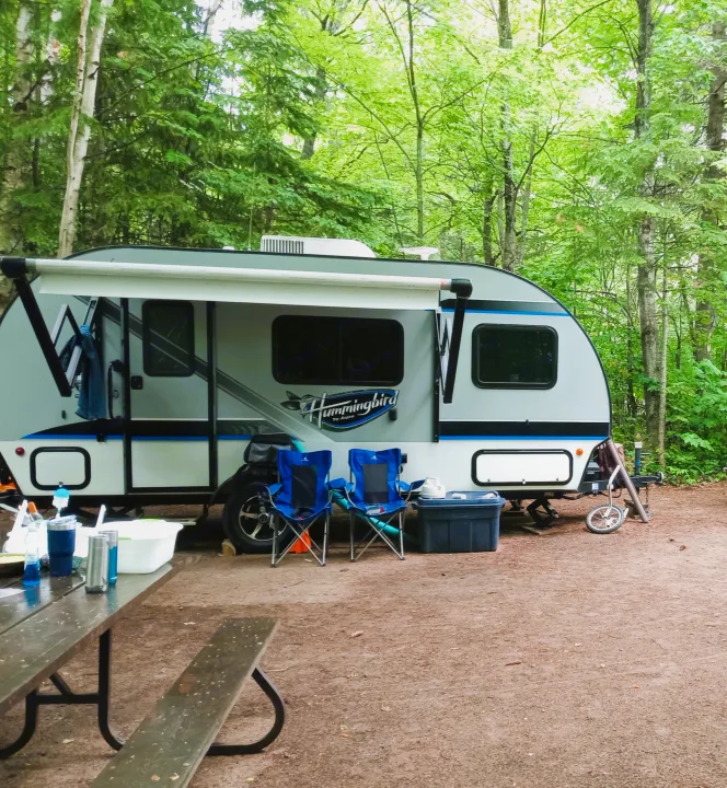A compact Jayco Hummingbird RV rental is set up in a lush green forest campsite, complete with blue chairs and a picnic table.. Jayco Hummingbird 2017
