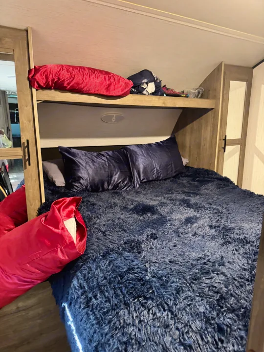 This RV rental features a cozy bunk bed setup: a dark blue fluffy lower bed with pillows, and an upper bunk with a red pillow.. Heartland RVs Pioneer 2022