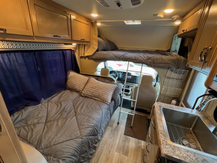 Spacious rv rental interior with a lower bed, upper bunk, kitchen sink, and driver's cockpit beyond. Ready for travel.. THOR MOTOR COACH Freedom Elite 2019