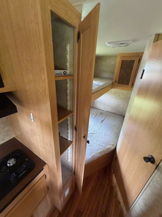 RV rental interior featuring wooden cabinetry, a compact kitchenette with a stove, open shelving, and bunk beds.. kz Coyote 2010