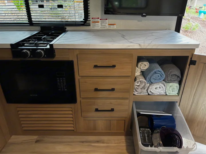 This RV rental's compact kitchen features a stove, microwave, drawers, and cabinet storage with towels and kitchenware.. Keystone Coleman Light 2026