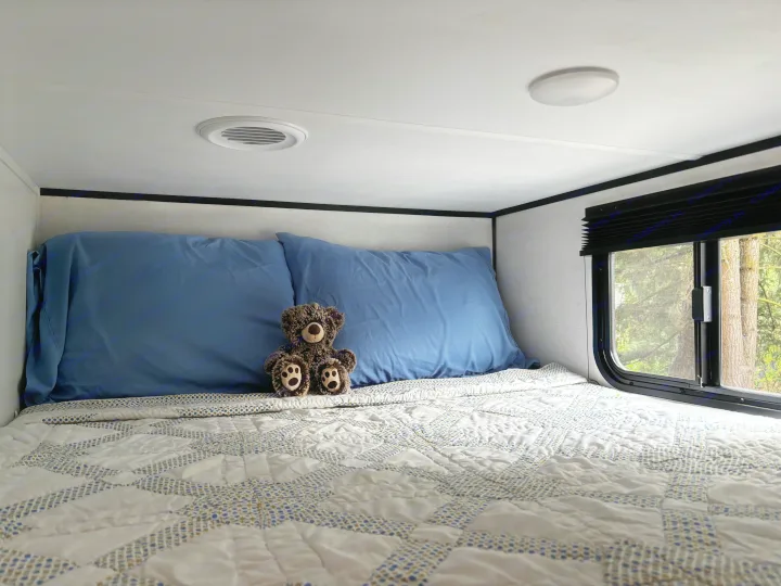 Cozy RV rental bed with blue pillows, patterned quilt, a teddy bear, and window view of trees.. Keystone Coleman Light 2026