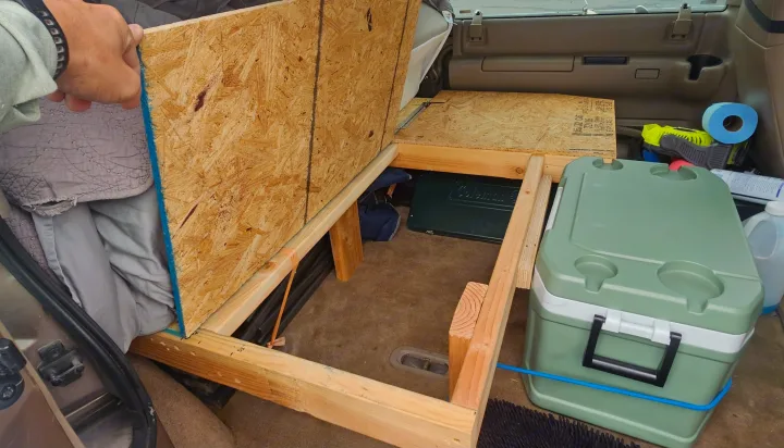 DIY car camping setup in a vehicle's rear. A wooden bed platform with storage, cooler, and stove offer an efficient rv rental alternative.. Chevrolet Astro 2003