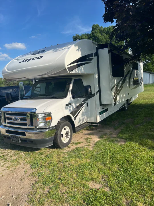 White Jayco RV rental, a spacious Class C motorhome, parked on a grassy lot under a clear blue sky. Adventure awaits!. Jayco Redhawk 2021