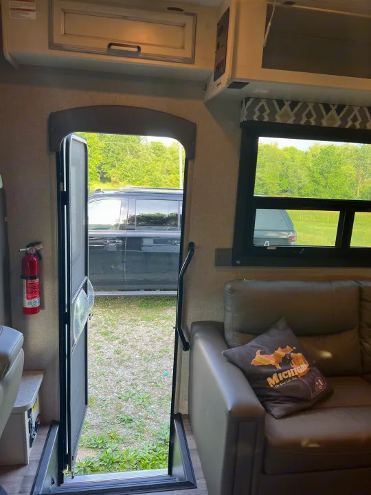 Inside an RV rental, looking out an open door with a screen, showing a parked SUV, trees, and gravel. Sofa with pillow nearby.. Jayco Redhawk 2021