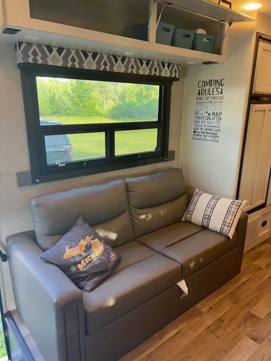 Cozy RV rental interior featuring a comfy grey sofa, scenic window view, overhead storage, and "Camping Rules" wall decal.. Jayco Redhawk 2021