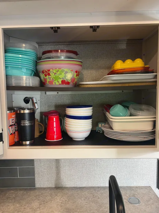 Organized RV rental kitchen cabinet with stacked plastic and ceramic dishes, bowls, and cups, showing smart storage.. Jayco Redhawk 2021
