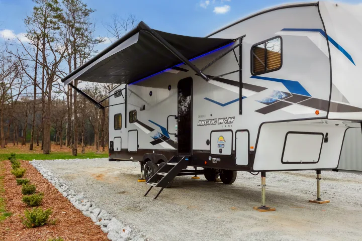 Modern fifth-wheel RV rental with illuminated awning, steps, and leveling jacks parked on a gravel lot amidst trees.. Cherokee limited Arctic Wolf 2023