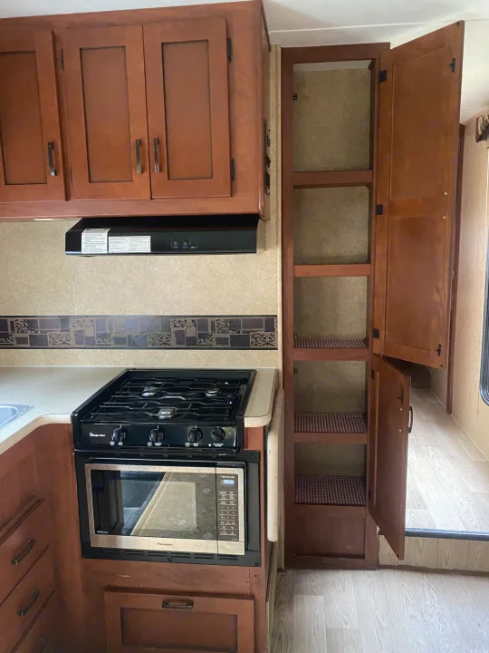 RV rental kitchen interior: features brown wood cabinets, gas stove, microwave, and an open pantry with multiple shelves.. Forest River Sunseeker 2011
