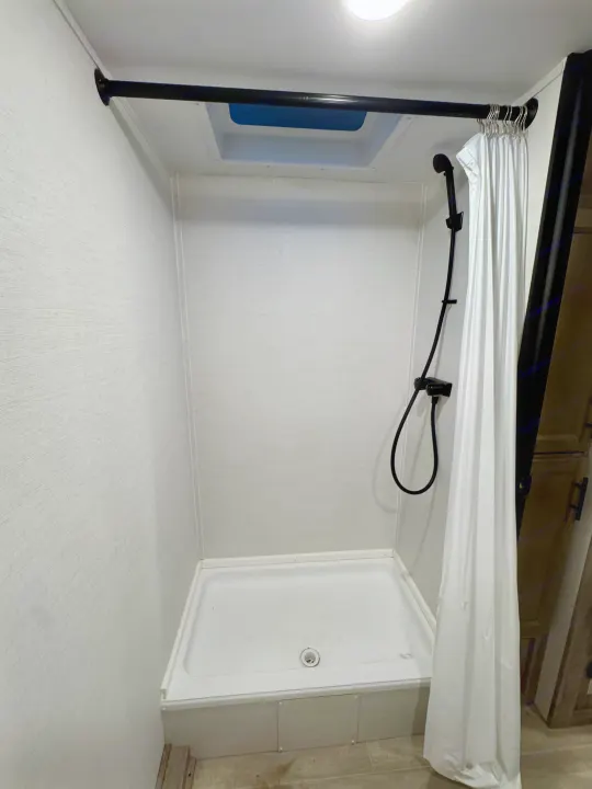 Compact shower in an RV rental, featuring a black shower head, white curtain, and ceiling vent for travel.. Gulf Stream Ameri-Lite 2025