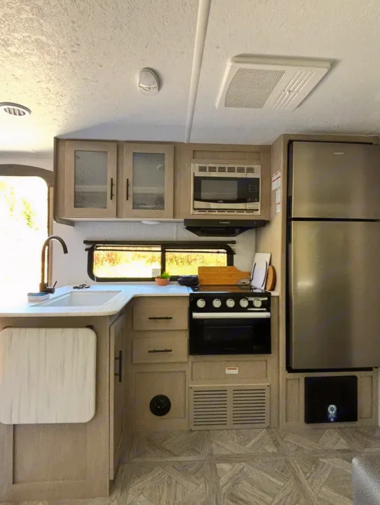 Inside an RV rental: a modern kitchen with stainless steel appliances, sink, light cabinetry, and wood-look flooring.. Forest River Wildwood X-Lite 2024