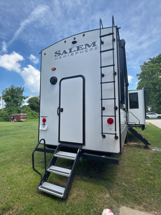 A white Salem Hemisphere travel trailer RV rental, with a rear ladder and side door steps on a green lawn under a blue sky.. Forest River Salem Hemisphere 2024