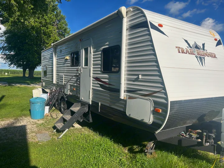 This white and gray Trail Runner RV rental is parked by a cornfield, showing its side entrance with steps, awning, and outdoor gear.. Heartland Trail Runner 2013