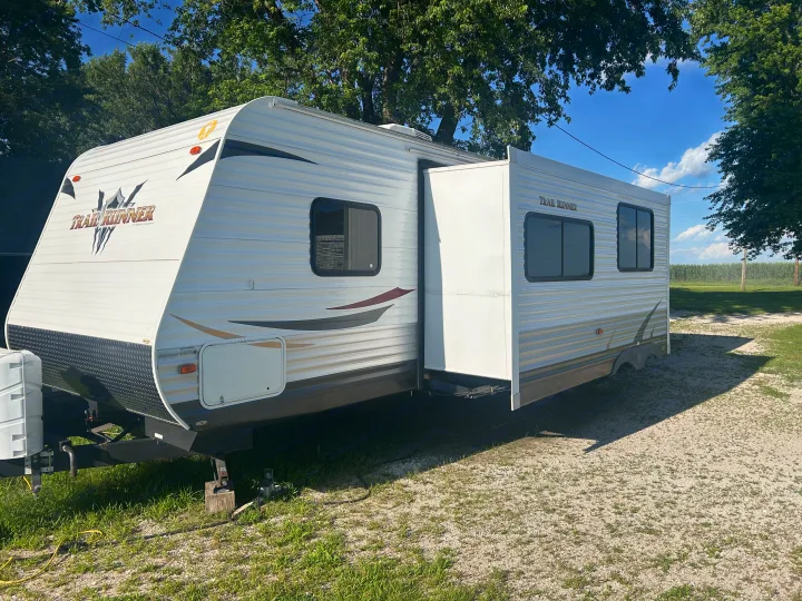 White Trail Runner RV rental with slide-out extended, parked on gravel. Blue sky & trees in the background.. Heartland Trail Runner 2013