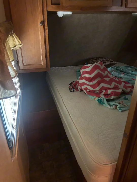 RV rental interior: A cozy bedroom with a white mattress, colorful blankets, wood cabinets, and a window with blinds.. Heartland Trail Runner 2013