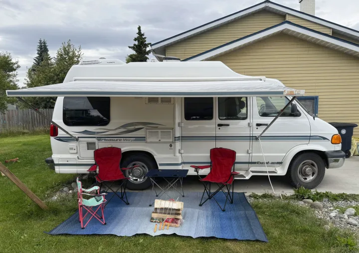 White RV rental with awning extended. Camping chairs, table, and firewood on a blue outdoor mat, ready for use.. Pleasure Way Excel 2001