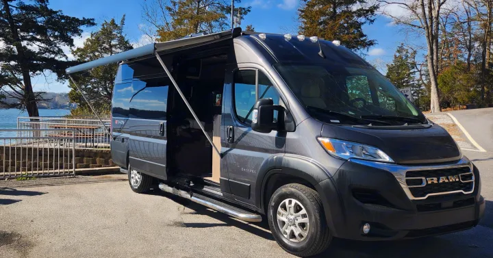 This grey Ram ProMaster rv rental is set up with its awning extended by a tranquil lake, ready for adventure.. Entegra Coach Ethos 2024