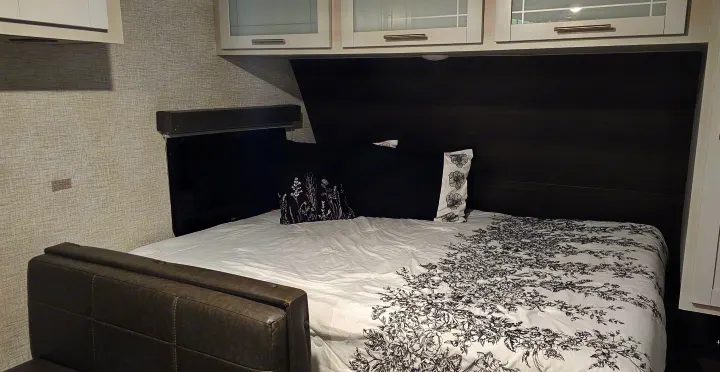 Cozy RV rental bedroom featuring a bed with elegant floral bedding, dark wood accents, and overhead cabinets.. Keystone RV Bullet Crossfire 2021