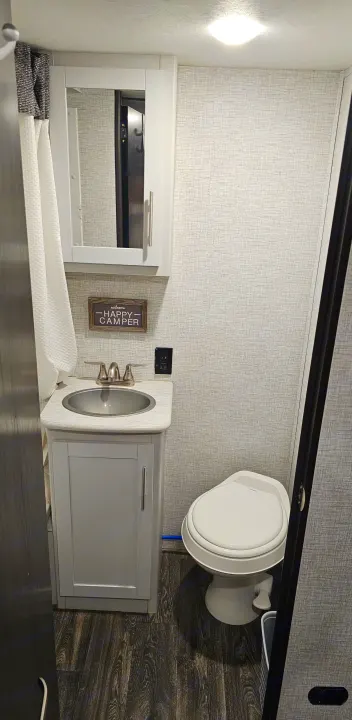 Inside an RV rental, this compact bathroom features a sink, toilet, mirror cabinet, and shower curtain. Textured walls, wood-look floor.. Keystone RV Bullet Crossfire 2021