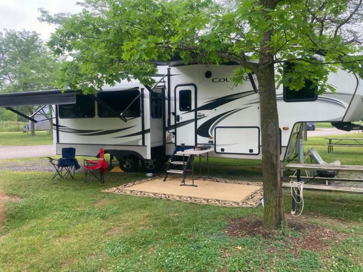 A white RV rental parked at a shady campsite. The awning is extended over an outdoor rug, chairs, and table.. Keystone RV Cougar 2020