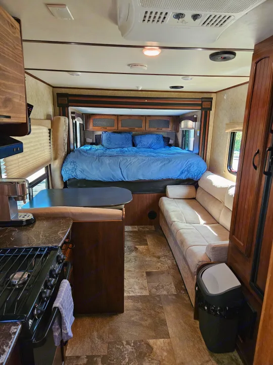 Bright RV rental interior. Features bedroom with blue bedding, comfy living room couch, & kitchen area with stove/coffee maker.. Jayco X213 2016