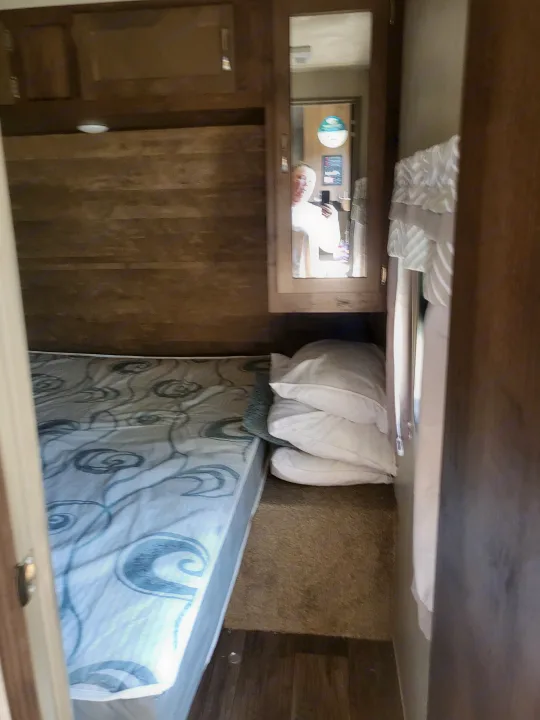 Compact sleeping area in an RV rental, with a patterned mattress, stacked pillows, and wooden overhead cabinets.. Forest River Palomino Puma 2018