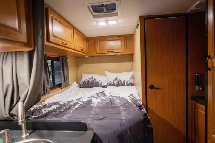Cozy RV rental interior features mountain-themed bedding, overhead cabinets, and a compact kitchen area with a sink.. Thor Majestic 2017