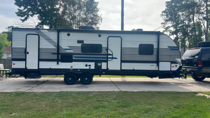 A modern gray and white travel trailer (RV rental option) is parked in a driveway, hitched to a black SUV.. Heartland RVs Pioneer 2023