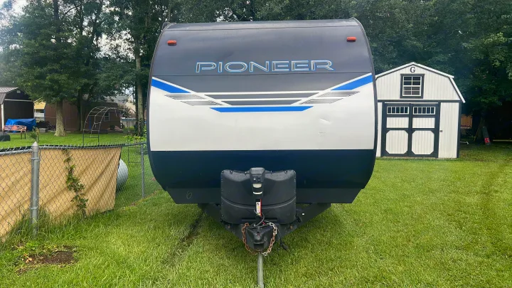 Front of a white and dark gray Pioneer travel trailer, ideal for rv rental. Parked on grass with sheds & trees visible.. Heartland RVs Pioneer 2023
