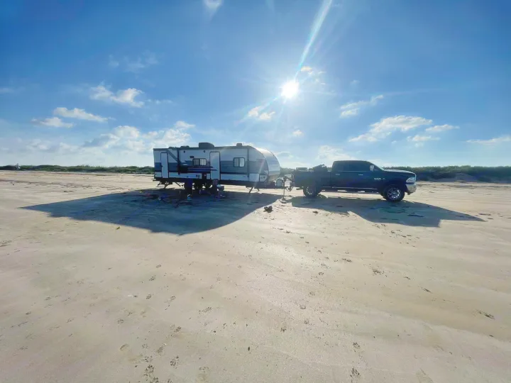 Experience an RV rental! Truck & travel trailer parked on a vast, sunny sandy beach under a clear blue sky.. Heartland RVs Pioneer 2023
