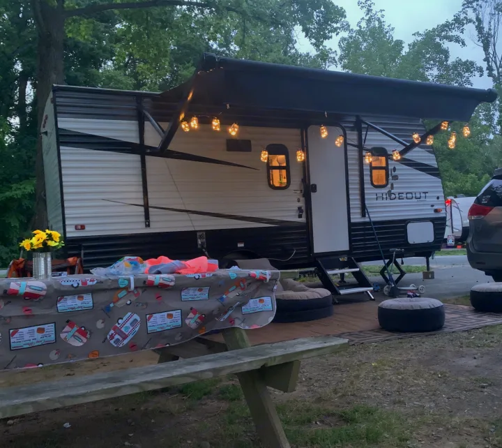 Relaxing rv rental campsite scene: travel trailer with illuminated awning, decorated picnic table, and comfy outdoor poufs.. Keystone RV Hideout 2021