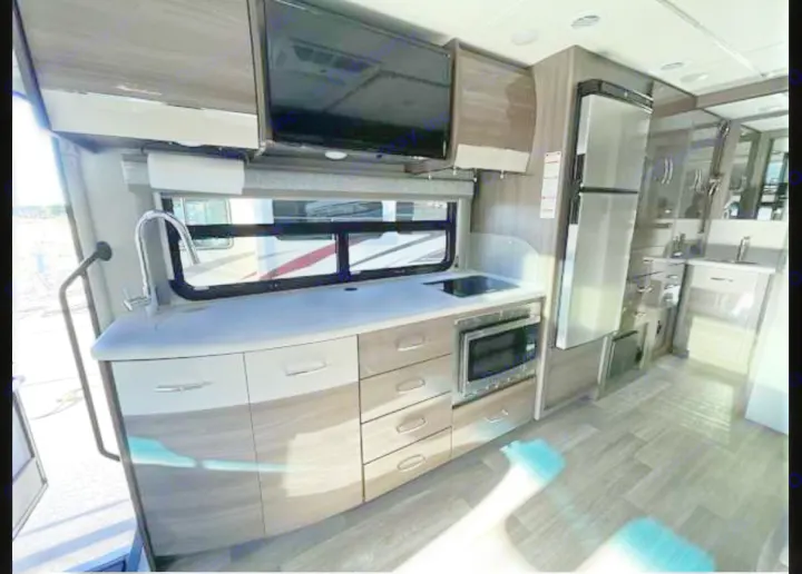 Inside a modern RV rental: a bright kitchen with sink, microwave, fridge, TV, and light wood cabinetry.. Thor Tiburon 2024