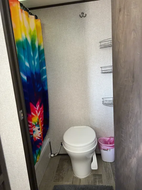 Compact RV rental bathroom: colorful tie-dye shower curtain, toilet, and wall-mounted shelves.. Dutchmen Coleman Lantern LT 2021