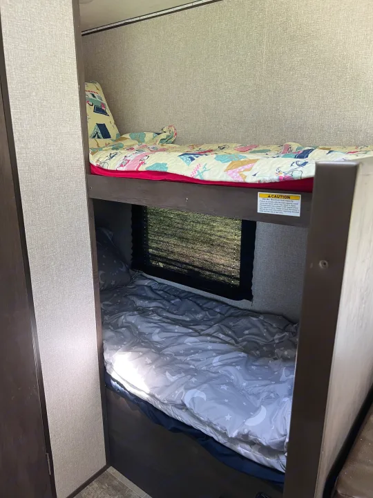 Inside an RV rental: functional bunk beds. Top bunk has a colorful quilt, bottom has moon/star bedding, with a window view. Ideal for travel.. Dutchmen Coleman Lantern LT 2021