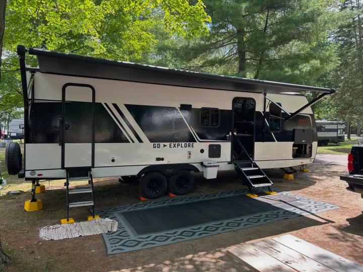 Modern RV rental set up at a shaded campsite with an extended awning, outdoor mat, and picnic table.. Cruiser RV x252 Go Explore 2025