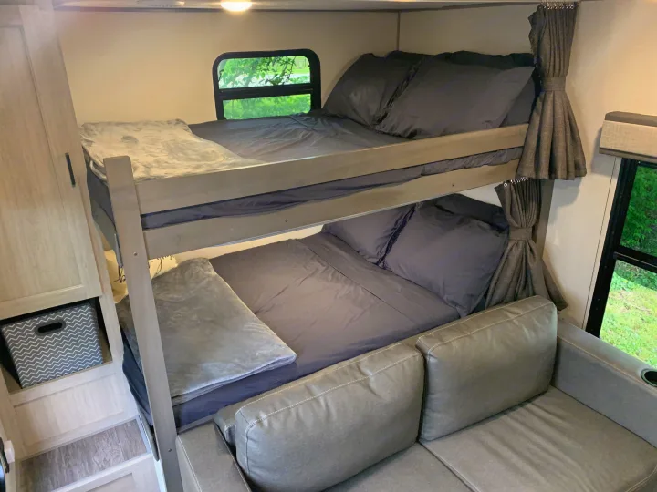 Cozy RV rental interior with gray bunk beds, a window showing greenery, a sofa, and storage. Perfect for family travel.. Forest River Aurora Aurora Light 2024