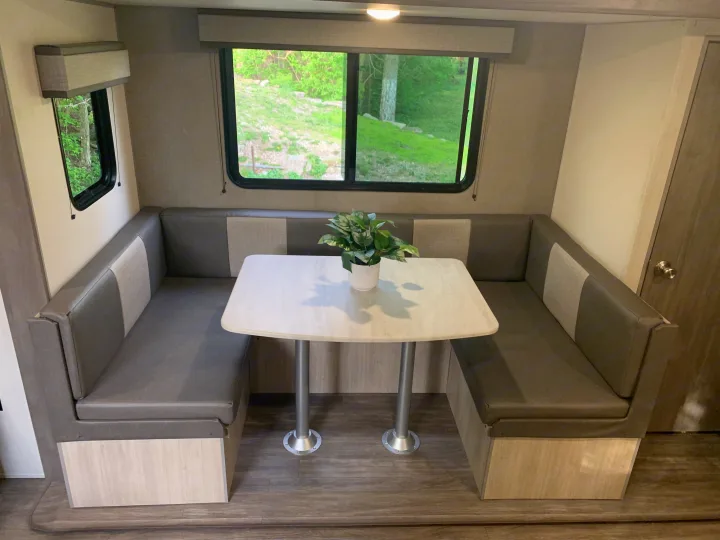 An RV rental dinette features U-shaped grey cushioned seating, a white table, and large windows revealing lush greenery outside.. Forest River Aurora Aurora Light 2024