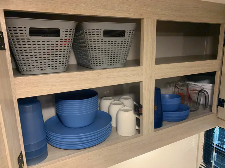 RV rental kitchen cabinet: organized blue dishes, white mugs, & storage baskets neatly stowed for easy travel.. Forest River Aurora Aurora Light 2024