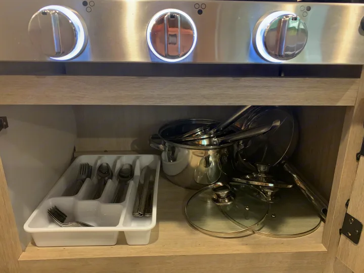 RV rental kitchen's open cabinet with organized silverware, stacked pots & pans, below a stove with illuminated knobs.. Forest River Aurora Aurora Light 2024